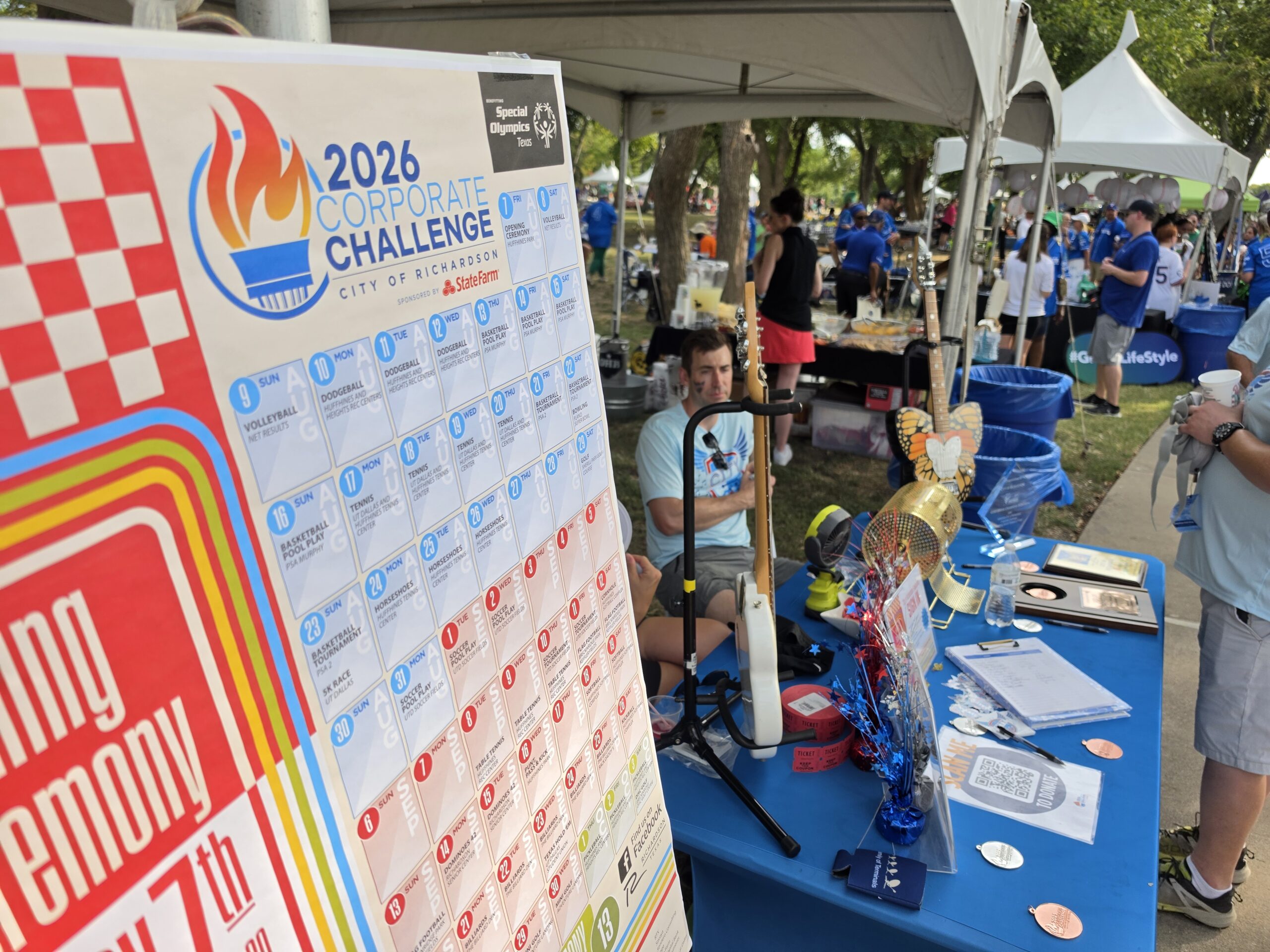 Booth at the 2026 City of Richardson Corporate Challenge featuring an event schedule, promotional materials, and attendees gathered under outdoor tents.